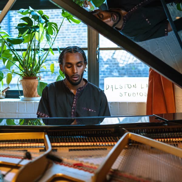 Judah at a grand piano with plants in the window behind, seen past the open lid