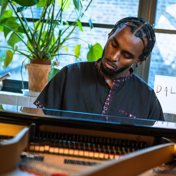 Judah playing a grand piano, plants in the window behind